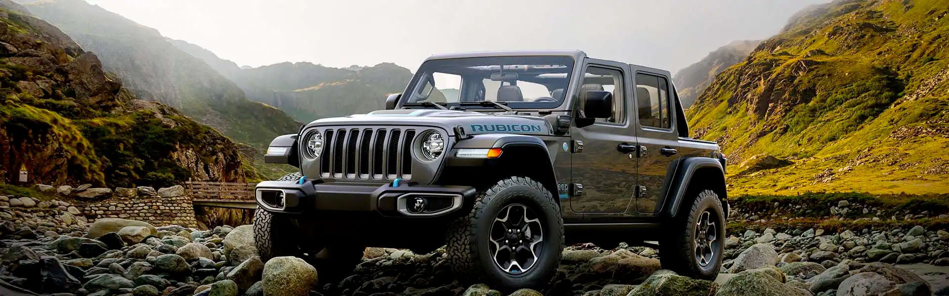 Hero image of the 2024 Jeep Wrangler 4xe parked on a rocky landscape in front of a stone bridge and a vast mountain landscape