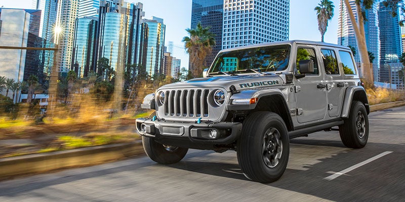 Image of the silver 2024 Jeep Wrangler 4xe Rubicon driving down a paved road lined with palm trees in a city
