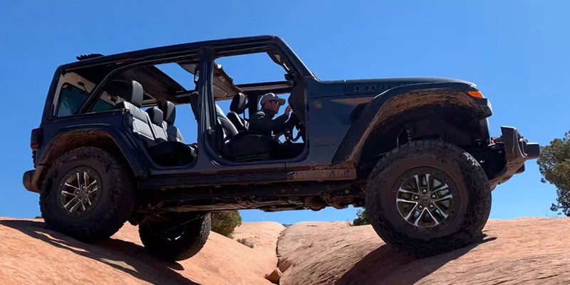Image of a black 2024 Jeep Wrangler shot at a lower angle to showcase its off-roading capabilities on uneven rock terrain