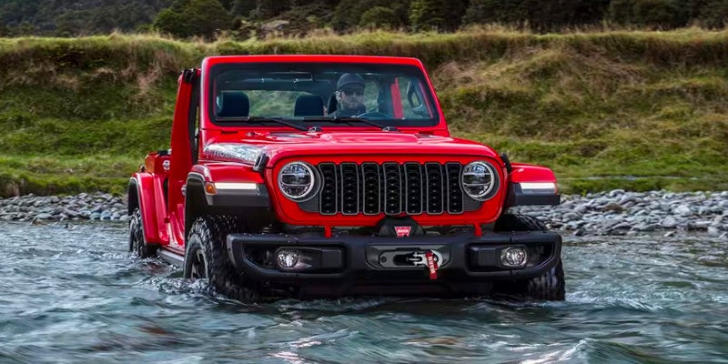 Image of a red 2024 Jeep Wrangler driving through a body of water with the tires halfway submerged, driving away from a grassy knoll