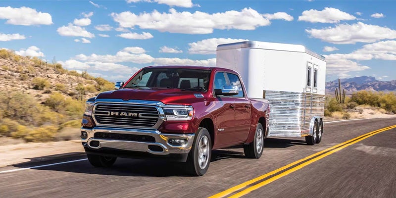 Image of a red 2024 RAM 1500 driving toward the camera on a highway in the desert, towing a white and chrome trailer