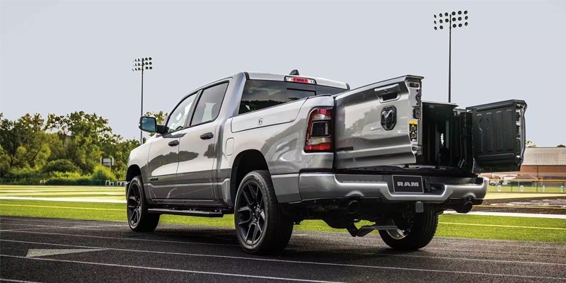 Image of a silver 2024 RAM 1500 facing away from the camera at an angle, parked on a running track of an outdoor sports facility, showcasing the multifunction tailgate doors