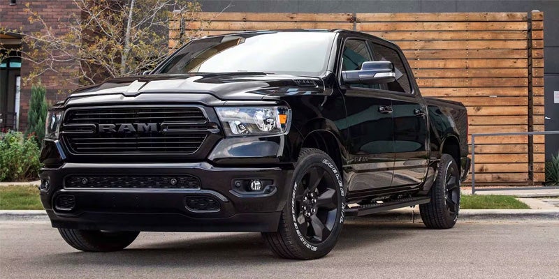 Image of a black 2024 RAM 1500 facing toward the camera at an angle, parked on a paved driveway in front of an urban home