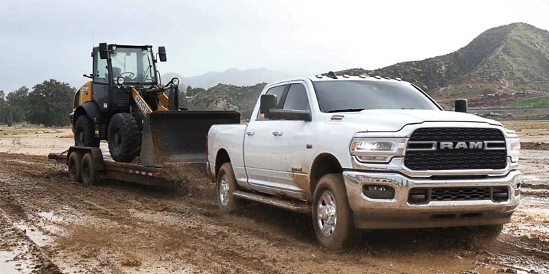 Image of the white RAM 2500 towing a bulldozer on a flatbed trailer through muddy terrain, with a mountainscape in the background