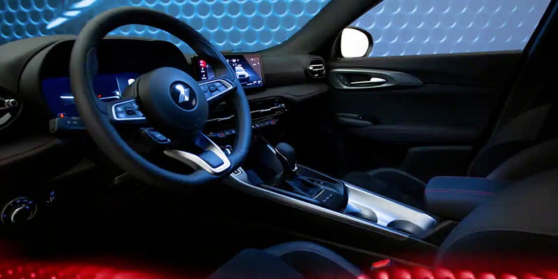 Image of the interior front dashboard of the 2024 Dodge Hornet, featuring black materials with red accent stitching on seats, infotainment system on and active, image shot from the driver's side