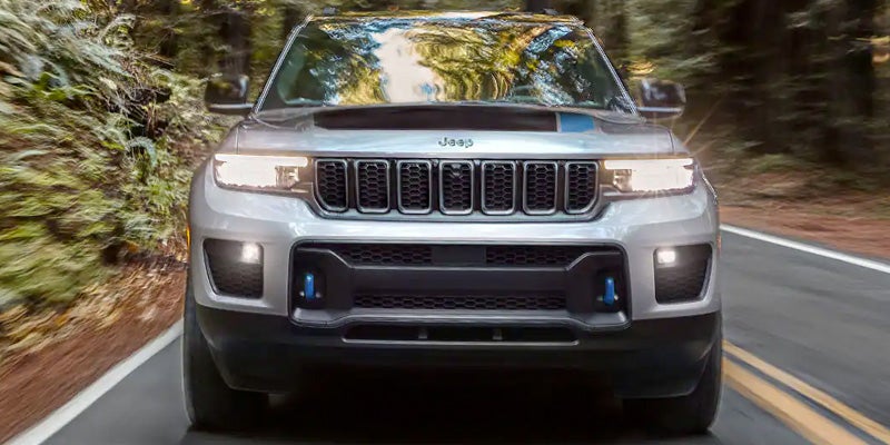 Image of the 2024 Jeep Grand Cherokee 4xe driving head-on down a two-lane road with trees blurring past on either side, displaying the Jeep seven-slot grille and headlights