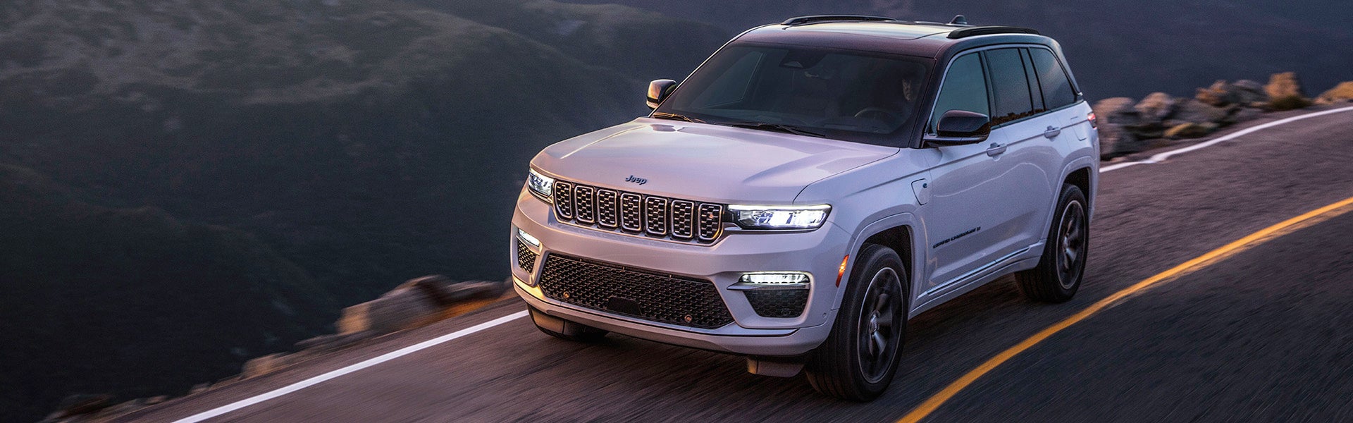 Hero image of a white 2024 Jeep Grand Cherokee driving on a highway through the mountains