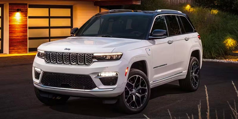 Image of a white 2024 Jeep Grand Cherokee posed at a front angle on a driveway in front of an urban home
