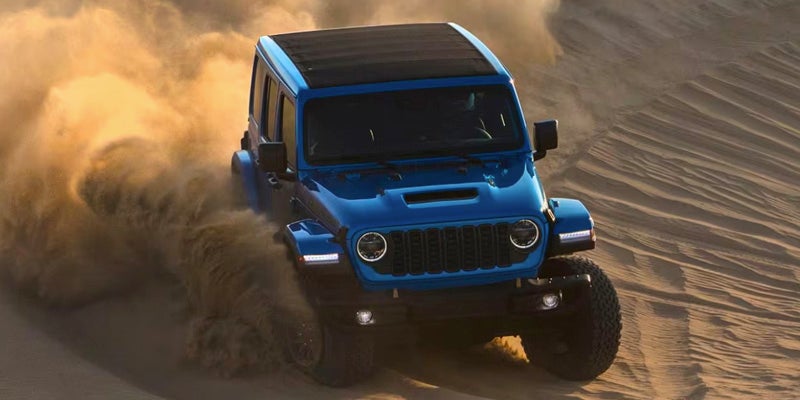 Image of a blue 2024 Jeep Wrangler shot in action from above, drifting through a desert dune and kicking up dust